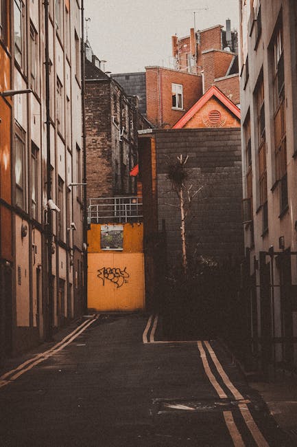 A narrow cobblestone city street in Whitechapel with tall brick and stone buildings on both sides. In the foreground, a red and white no entry traffic sign is mounted on a black pole, positioned on th
