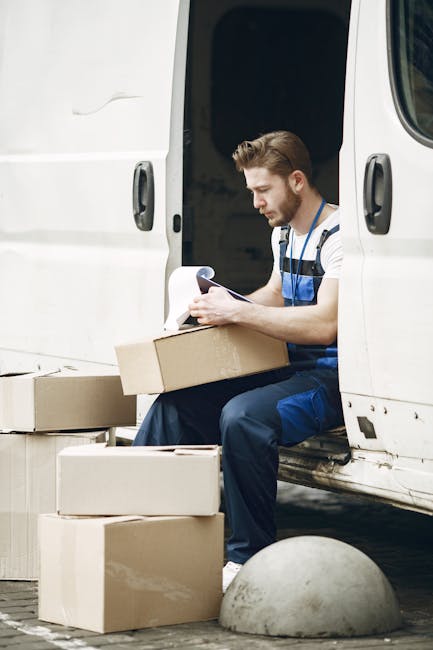 A man dressed in a blue jacket and dark trousers is loading or unloading a cardboard box from the open cargo area of a white panel van parked on a city street. The van, which features an empty interio