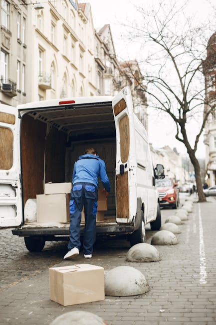 A large green box truck with a closed cargo area is parked on a city street, facing away from the camera, with its rear doors visible. The truck is positioned near the curb in front of modern high-ris