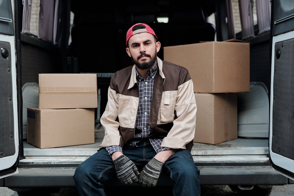 A man wearing a white shirt, jeans, and a black cap is seen unloading cardboard boxes from a white panel van parked on a residential street. The man is carrying a stack of three medium-sized cardboard