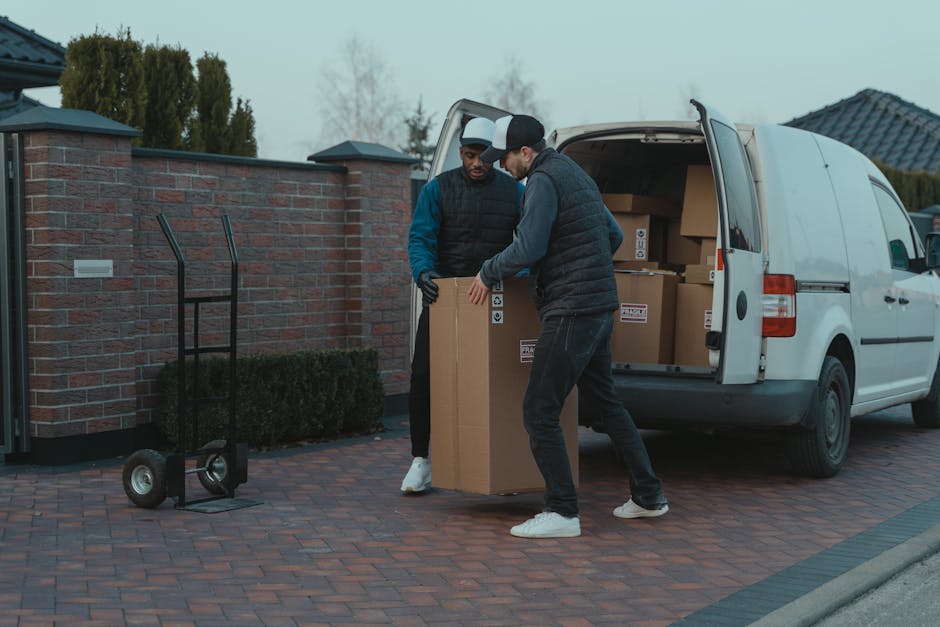Two men engaged in a home relocation process are loading a large cardboard box into the back of a white van parked outside a residential property. The van’s rear doors are open, revealing several other cardboard boxes of various sizes stacked inside. One man, wearing a black quilted vest over a grey hoodie and white sneakers, is lifting and guiding the box into the van, while the other, dressed in a navy jacket with blue sleeves and black pants, assists in positioning the box. The scene takes place on a paved driveway bordered by a brick wall and well-maintained shrubbery, with a two-storey house visible in the background under an overcast sky. An empty hand truck with two wheels and a vertical frame is standing nearby, ready for further use in the packing and moving operation, which is part of a professional removals service by Man and Van Whitechapel specializing in furniture transport and domestic relocations.