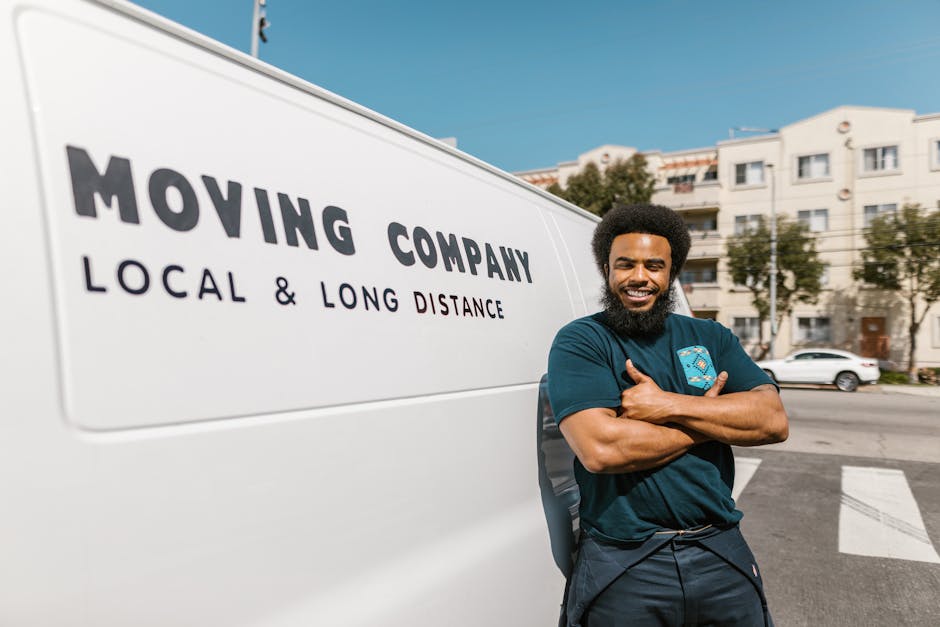 A smiling man with a dark beard and afro hairstyle stands in front of a white moving van marked with the text 'MOVING COMPANY LOCAL & LONG DISTANCE'. He is wearing a navy T-shirt with a logo on the chest, arms crossed, and appears to be outside a residential area with apartment buildings and parked cars under a clear blue sky. The scene depicts the loading or transportation process of home relocation, with the van positioned on the street near the pavement, indicating a local or long-distance furniture transport service. The background includes trees and modern white buildings, and the lighting suggests daylight, emphasizing the professional setting of a house or apartment move. This image illustrates the services of [COMPANY_NAME], providing professional removals and packing assistance as part of house moving logistics.