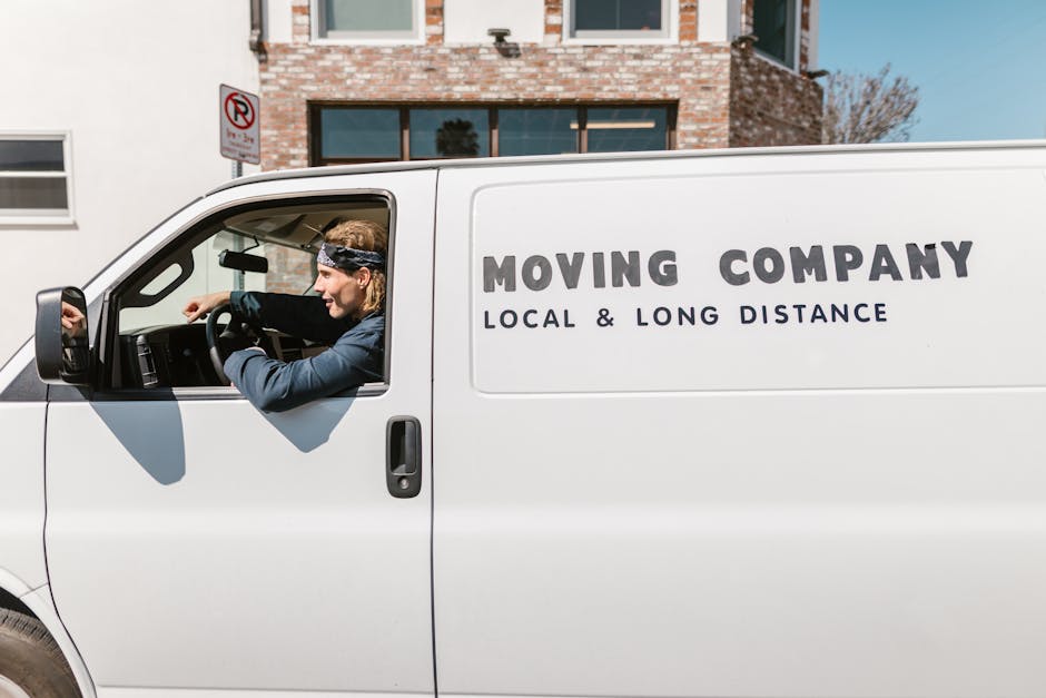 Two men engaged in a home relocation process are loading a large cardboard box into the back of a white van parked outside a residential property. The van’s rear doors are open, revealing several other cardboard boxes of various sizes stacked inside. One man, wearing a black quilted vest over a grey hoodie and white sneakers, is lifting and guiding the box into the van, while the other, dressed in a navy jacket with blue sleeves and black pants, assists in positioning the box. The scene takes place on a paved driveway bordered by a brick wall and well-maintained shrubbery, with a two-storey house visible in the background under an overcast sky. An empty hand truck with two wheels and a vertical frame is standing nearby, ready for further use in the packing and moving operation, which is part of a professional removals service by Man and Van Whitechapel specializing in furniture transport and domestic relocations.