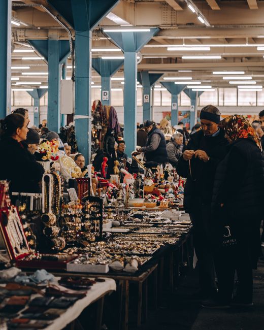 Inside a large industrial market hall, featuring blue steel support columns and bright overhead lighting, with numerous vendors and shoppers browsing tables filled with an array of jewelry, accessorie