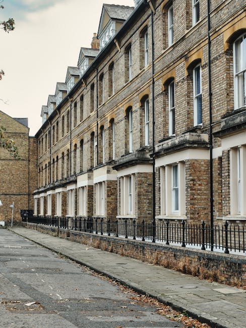 A row of older brick terraced houses with white-framed sash windows and small front gardens enclosed by black wrought iron fences, situated along a narrow paved street in Whitechapel. The pavement is lined with concrete slabs, and some fallen leaves are scattered on the ground. The sky is overcast, casting soft diffuse light over the scene. This residential street is typical for house removals and home relocation services, with potential logistics involving furniture transport through narrow pathways. The image is associated with the Whitechapel removals guide, highlighting the typical environment for moving services provided by Man and Van Whitechapel. There are no people or vehicles visible, focusing solely on the exterior of the historic housing stock.