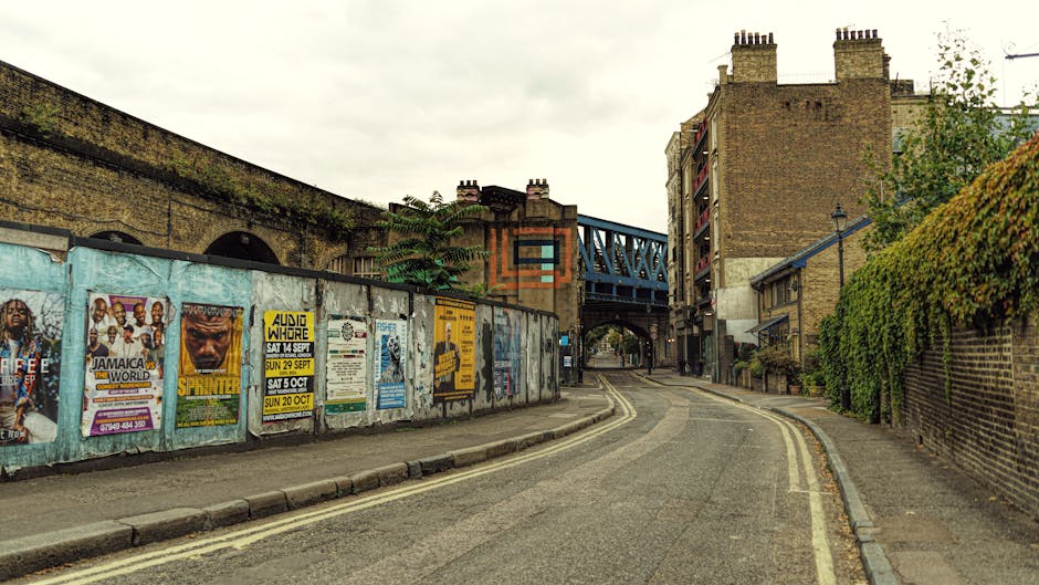 A narrow, slightly curved street in Whitechapel featuring a residential and commercial urban environment with a cloudy sky overhead. On the left side, a weathered metal fence covered in colourful posters advertising events and music gigs separates the sidewalk from an elevated brick structure, possibly part of a railway or bridge. The sidewalk is paved with concrete, with some visible wear, and a small curb runs along the edge. To the right, a tall brick wall covered in dense, green foliage borders a property, with a lamppost positioned at the fence corner. In the background, there is a blue metal bridge spanning over the road, connecting the upper parts of the buildings, which include brick apartments with multiple floors and chimneys on the rooftops. The street appears to be part of a typical urban house removal or furniture transport route, supported by the surrounding environment consistent with city moving and packing logistics, as would be seen during house removals in Whitechapel. Man and Van Whitechapel's service involves loading furniture, boxes, and appliances onto vehicles in this area for home relocation purposes.