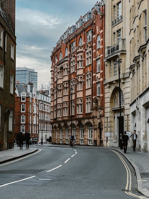 A row of older brick terraced houses with white-framed sash windows and small front gardens enclosed by black wrought iron fences, situated along a narrow paved street in Whitechapel. The pavement is lined with concrete slabs, and some fallen leaves are scattered on the ground. The sky is overcast, casting soft diffuse light over the scene. This residential street is typical for house removals and home relocation services, with potential logistics involving furniture transport through narrow pathways. The image is associated with the Whitechapel removals guide, highlighting the typical environment for moving services provided by Man and Van Whitechapel. There are no people or vehicles visible, focusing solely on the exterior of the historic housing stock.